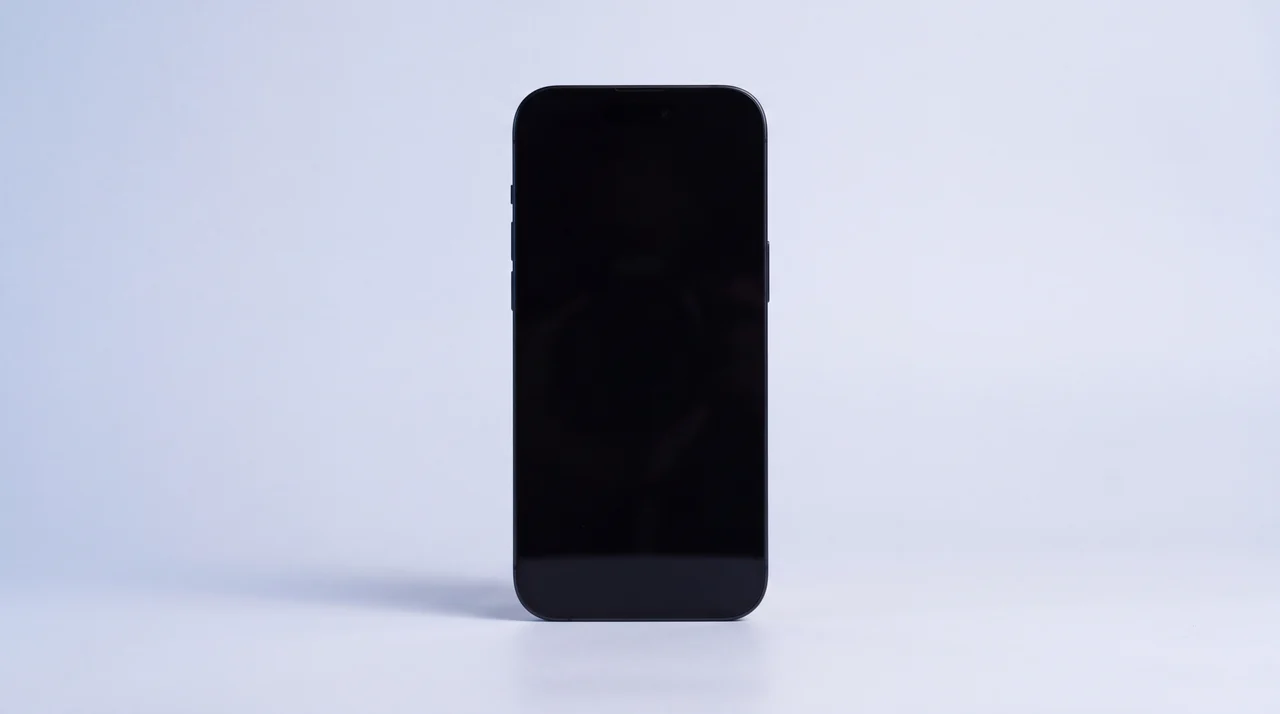 A single pristine black iPhone 15 standing upright on a pale grey surface with its screen dark, nothing else in frame