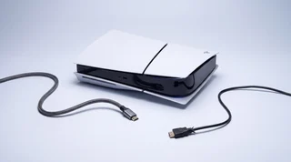 A PlayStation 5 standing upright on a clean pale-grey studio surface with an HDMI cable connected to the rear port and a subtle blue glow along the cable