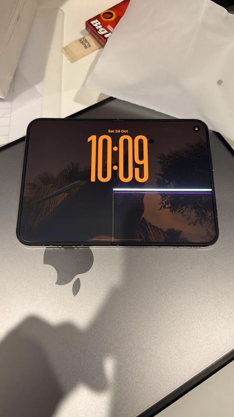 An open iPhone Duo on a desk showing a lock screen, with a dark point at the hinge, a thin vertical line running down the crease and a bright horizontal band across the right-hand half