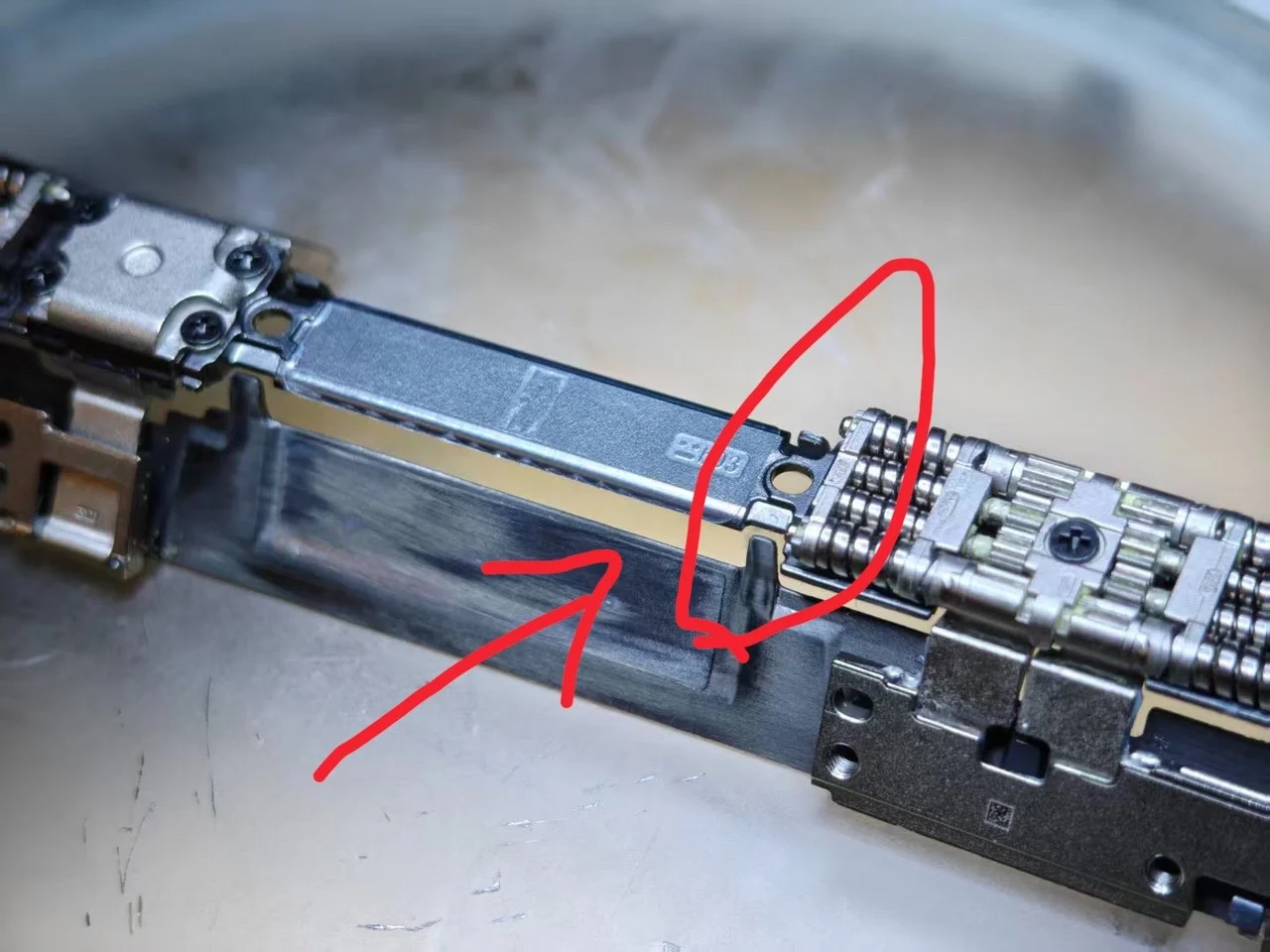 A disassembled iPhone Duo hinge on a bench: the long centre plate with a tab at its end circled in red beside the hinge gear block, and a red arrow pointing at a bright rub mark on the plate next to it