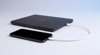 A black iPhone 15 lying face up beside a closed MacBook Pro on a pale grey bench, joined by a single USB-C cable into the bottom of the phone, ready for a full erase and reinstall