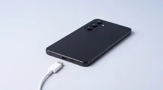 A black Samsung Galaxy S25 lying face down on a pale-grey surface with a white USB-C cable unplugged just in front of its charging port