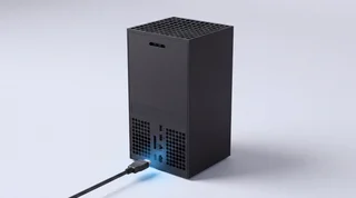 A single Xbox Series X console standing upright on a clean pale-grey studio surface with an HDMI cable connected at the base and a subtle blue glow at the connection point